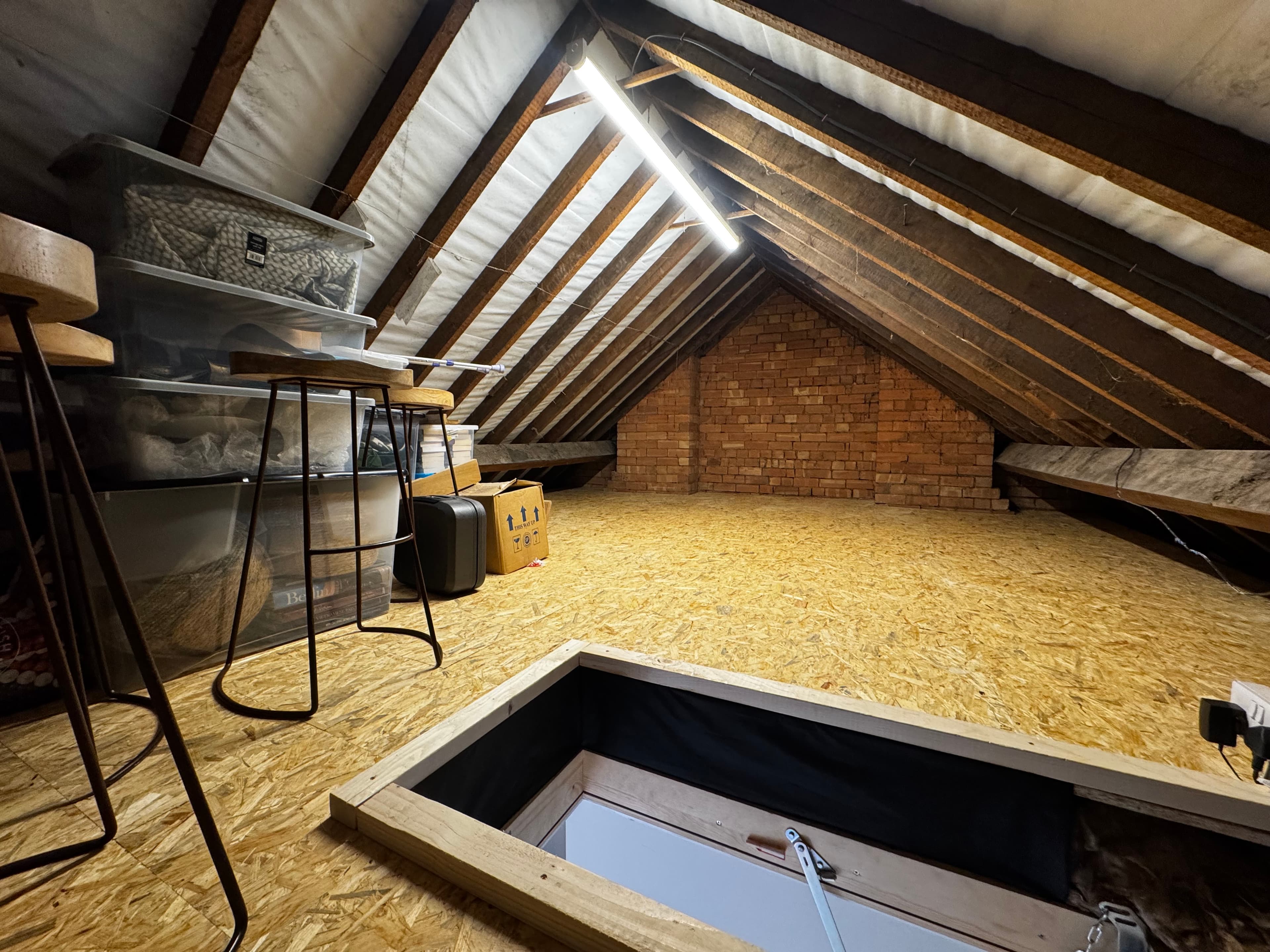 Bright attic with wooden rafters, OSB flooring, storage bins, and an open floor hatch.