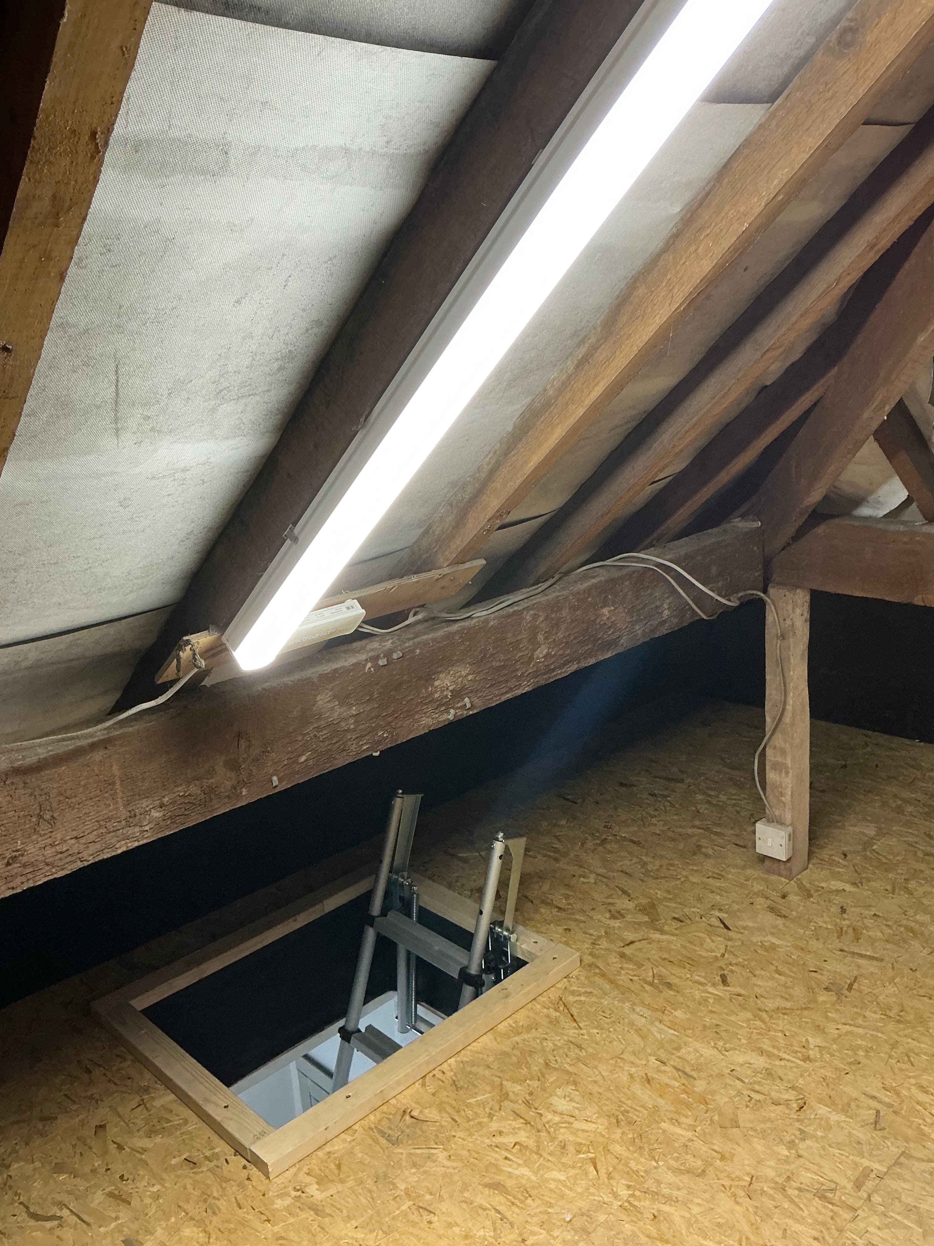 Bright light illuminates an attic with wooden rafters, OSB flooring, and an open hatch ladder.
