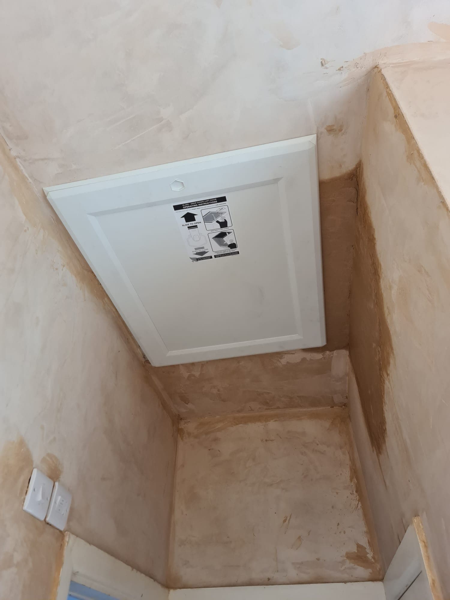 White loft access hatch installed in a ceiling surrounded by unfinished, freshly plastered brown walls.