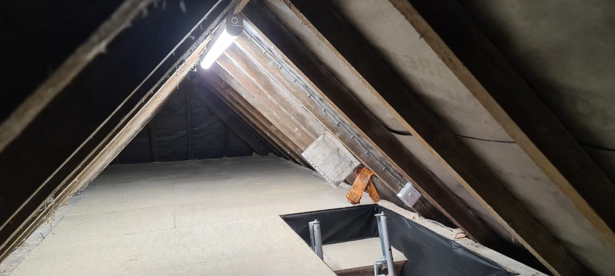 Brightly lit boarded attic space with slanted wooden rafters and an open loft hatch.