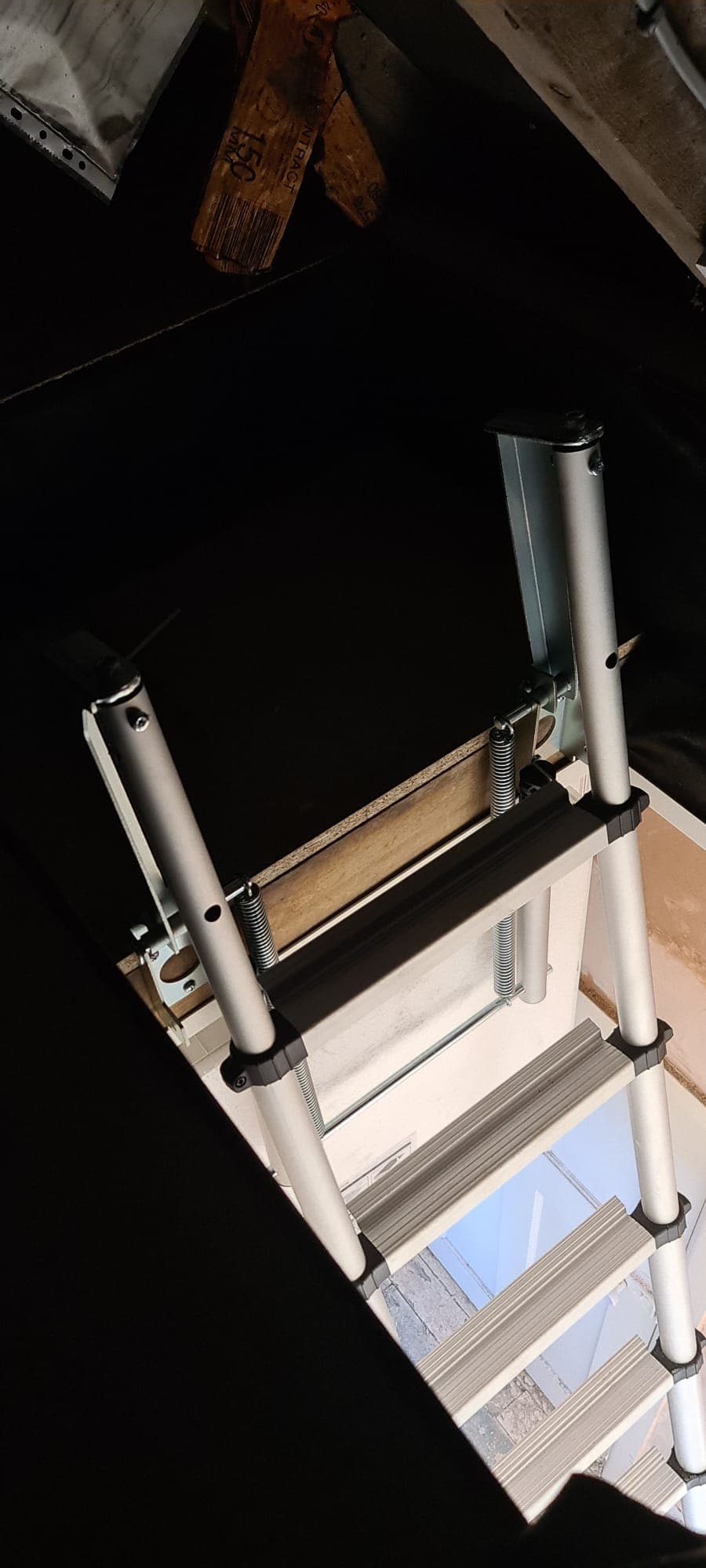 Silver telescopic loft ladder extending from a dark attic into a brightly lit room below.