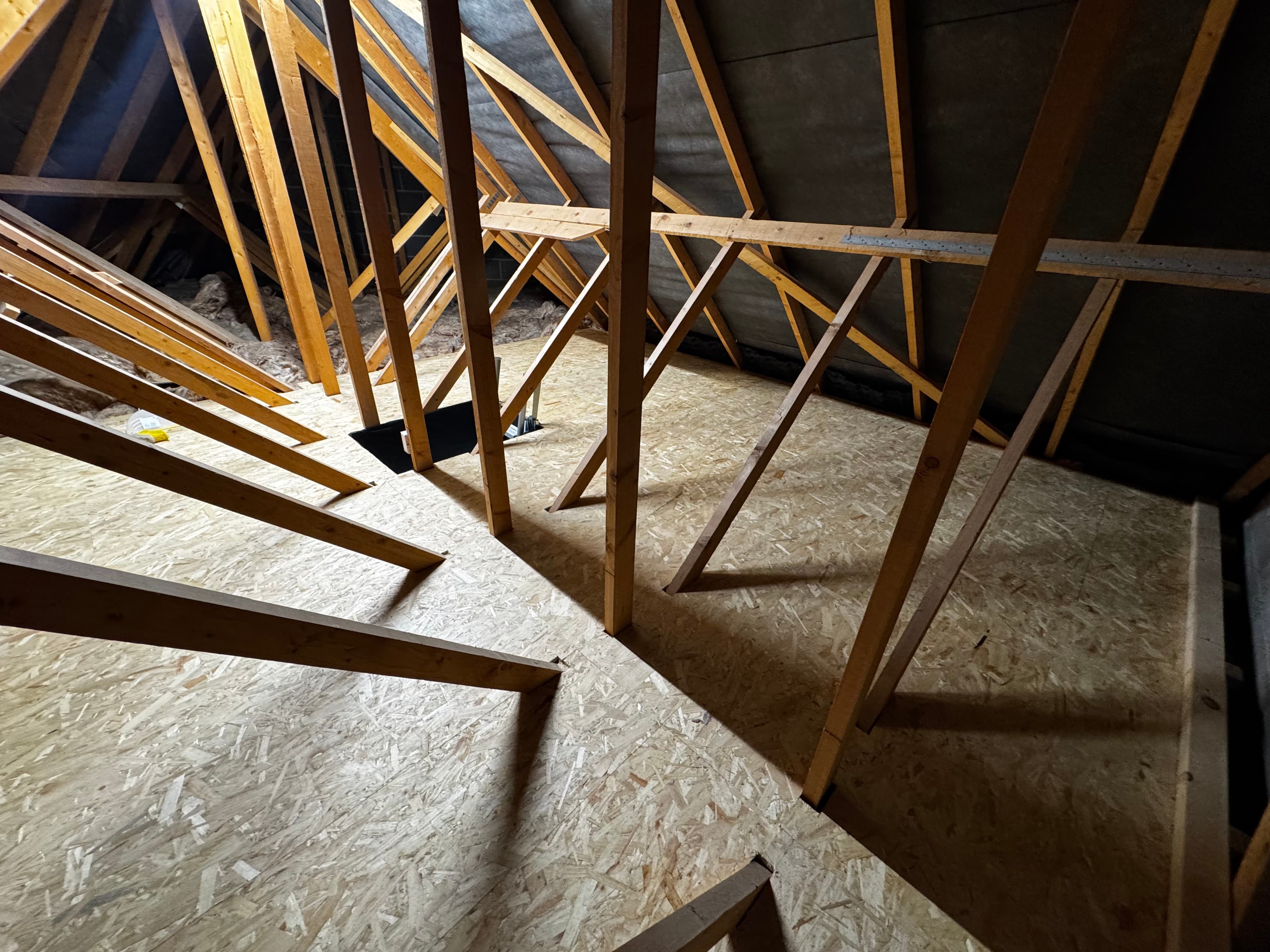 Unfinished attic with wooden roof trusses, OSB flooring, and a dark rectangular access hatch.