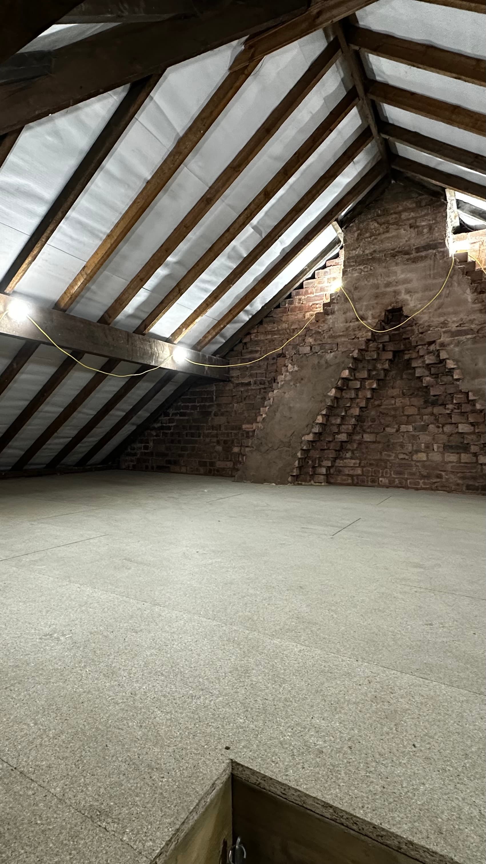 Unfinished attic featuring exposed wooden rafters, chipboard flooring, and an old red brick wall.